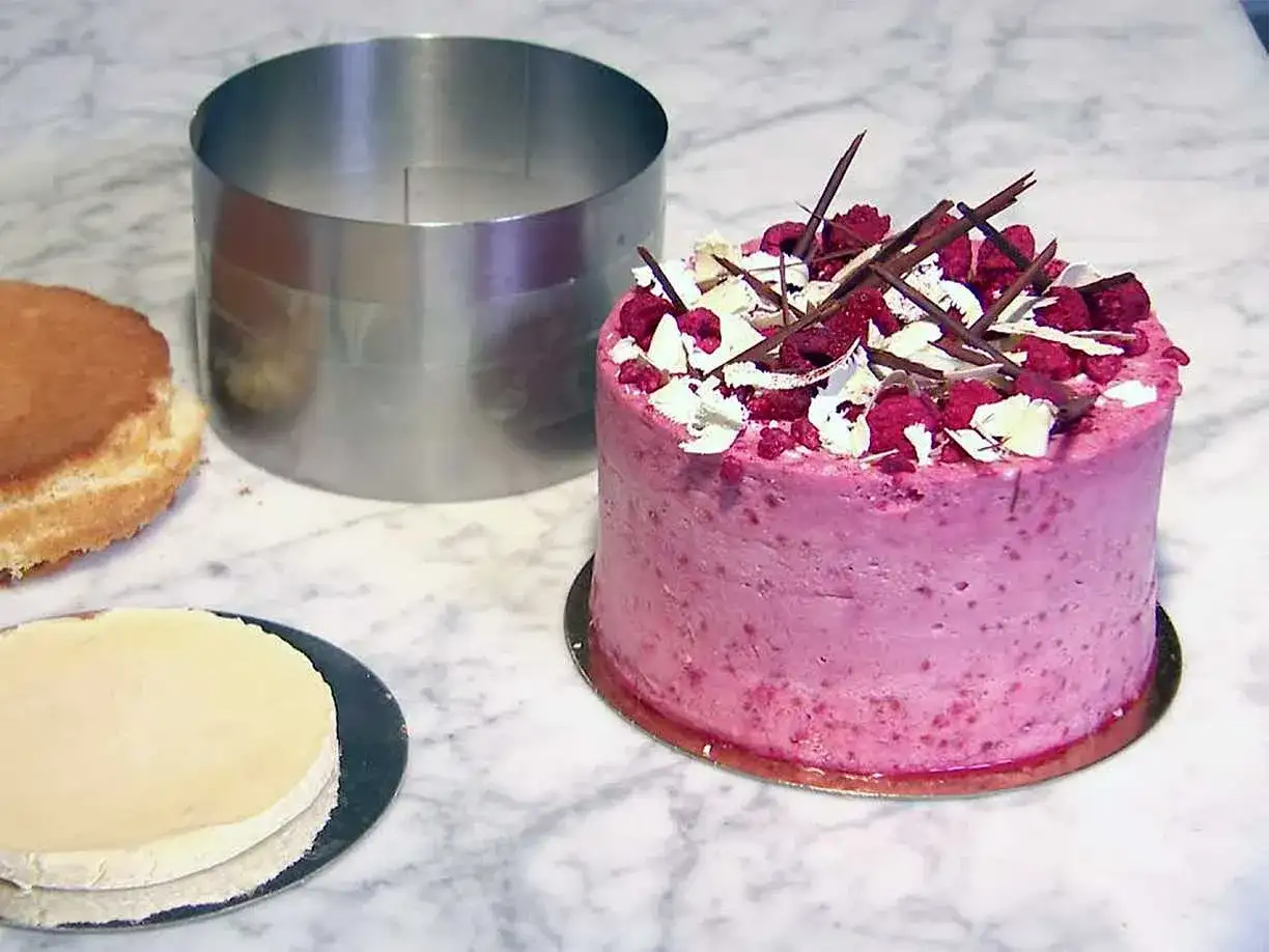 En rosa hallontårta med choklad och vita dekorationer. Bredvid ligger en bakring och delar av en tårta, redo för ett nytt tårta recept.