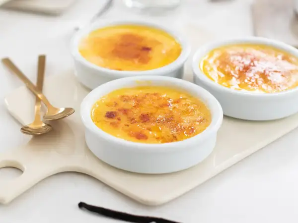 Tre krämiga creme brulee i vita skålar med karamelliserat sockerlock. Guldfärgade skedar ligger bredvid.