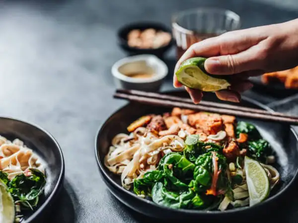 En hand pressar lime över en skål med nudlar och grönsaker, perfekt vin till asiatisk mat.