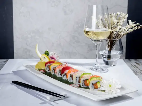 En elegant uppsättning sushirullar med färgglada toppingar, serverade med en glas vin till sushi. En vacker dukning med blommor och ätpinnar.