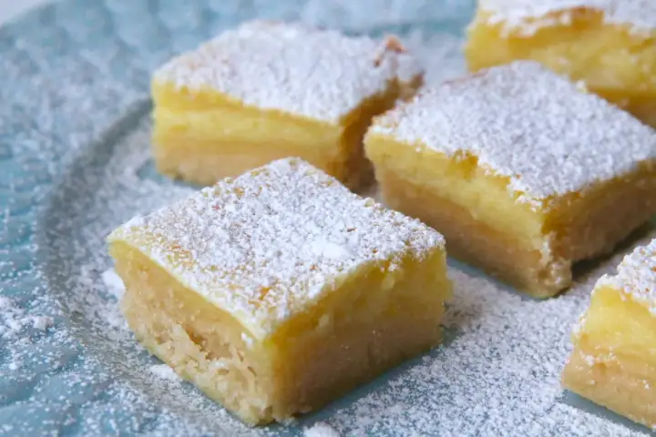 Färska bitar av citronkaka i långpanna, pudrade med florsocker, ligger på en blå tallrik.